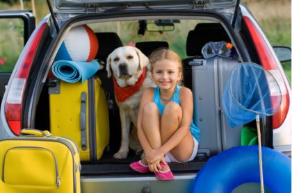 young girl and her dog in the back of a car packed for a summer trip